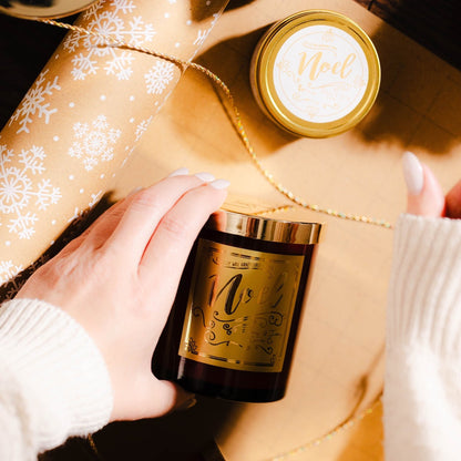 A red 8oz soy candle with a gold lid and a gold label named "noel" being wrapping in brown and white snowflake wrapping paper and gold twine.