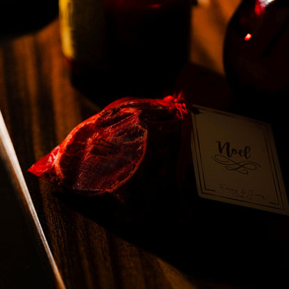 A small red organza bag with a white hang tag named "noel" filled with dried botanicals, sitting on a wooden table.