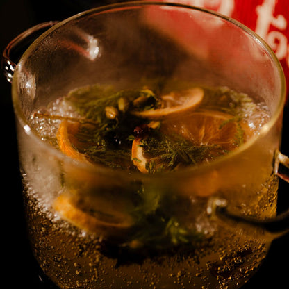 A clear simmer pot simmering on a stone with dried oranges, lemons, cedar branches, cinnamon, and cloves.