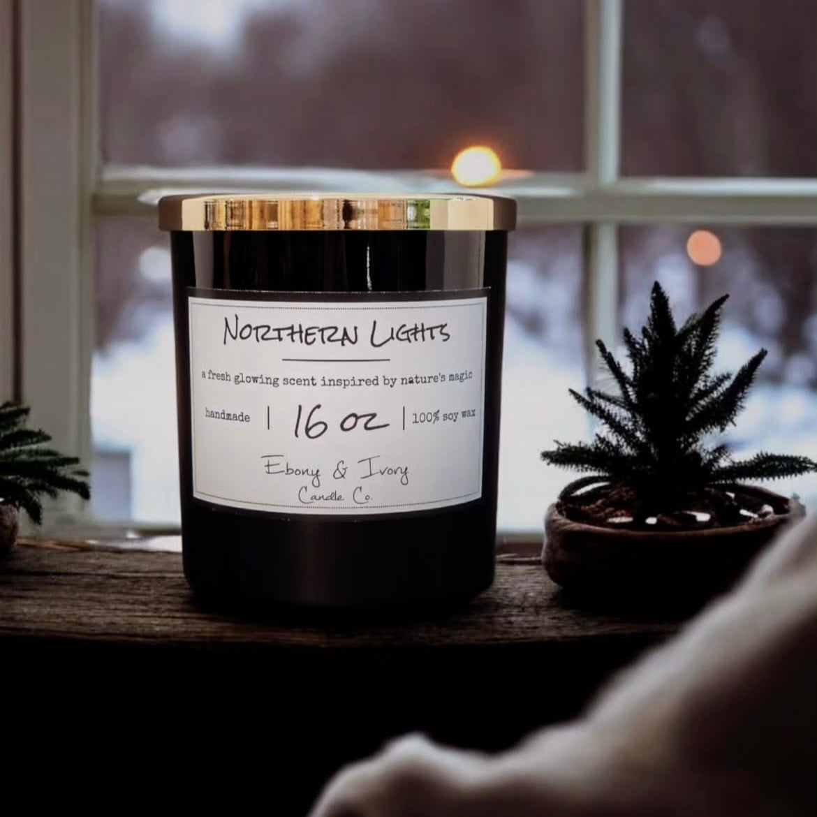 A black jar 16oz soy candle with a gold lid and a white label named Northern Lights, in a winter window sill, made by Ebony & Ivory Candle Co in Lethbridge, Alberta, Canada