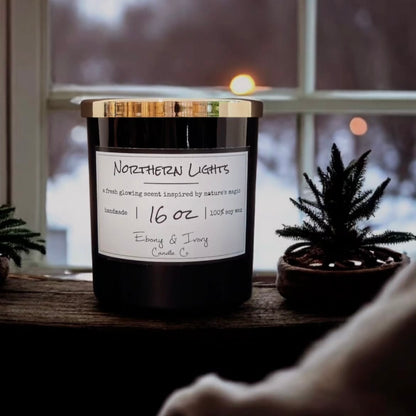 A black jar 16oz soy candle with a gold lid and a white label named Northern Lights, in a winter window sill, made by Ebony & Ivory Candle Co in Lethbridge, Alberta, Canada
