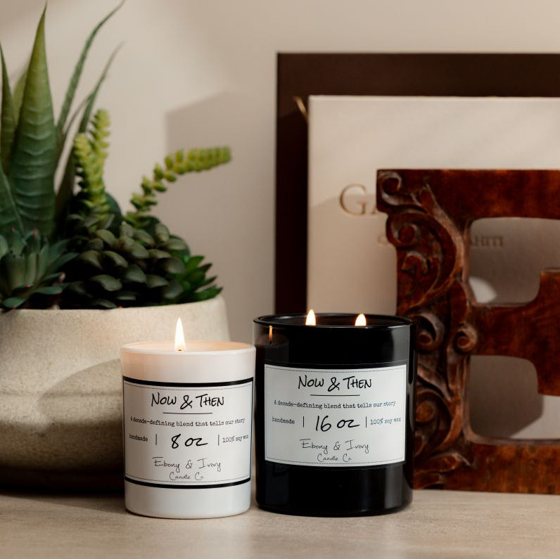 a Black 16oz soy candle with two cotton wicks and a white 8oz soy candle with one cotton wick, sitting on a neutral background with a plant, two arts books, and a decorative wooden 'E'. The candle is labelled Now & Then, made by ebony & ivory candle co.
