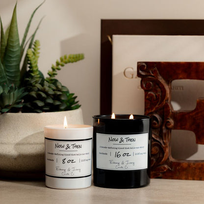 a Black 16oz soy candle with two cotton wicks and a white 8oz soy candle with one cotton wick, sitting on a neutral background with a plant, two arts books, and a decorative wooden 'E'. The candle is labelled Now & Then, made by ebony & ivory candle co.