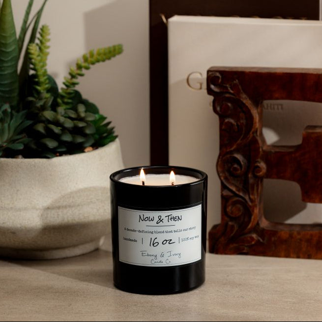 a Black 16oz soy candle with two cotton wicks sitting on a neutral background with a plant, two arts books, and a decorative wooden 'E'. The candle is labelled Now & Then, made by ebony & ivory candle co.