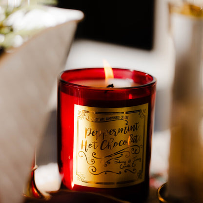 A red 8oz soy christmas candle with a gold label named "peppermint hot chocolate" sitting on a christmas dining table.