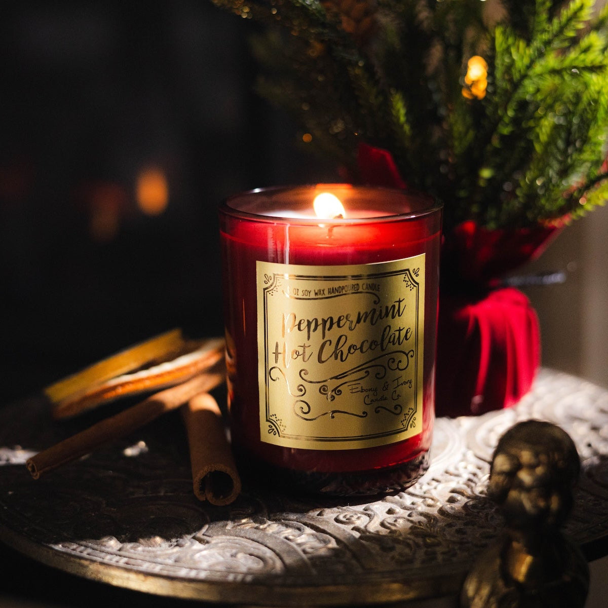 A red 8oz soy christmas candle with a gold label named "peppermint hot chocolate" sitting on a gold side table with cinnamon sticks and christmas decorations