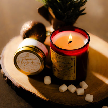 A red 8oz soy christmas candle with a gold label named "peppermint hot chocolate" sitting next to a gold 4oz soy candle on a tree cookie with marshmallows.
