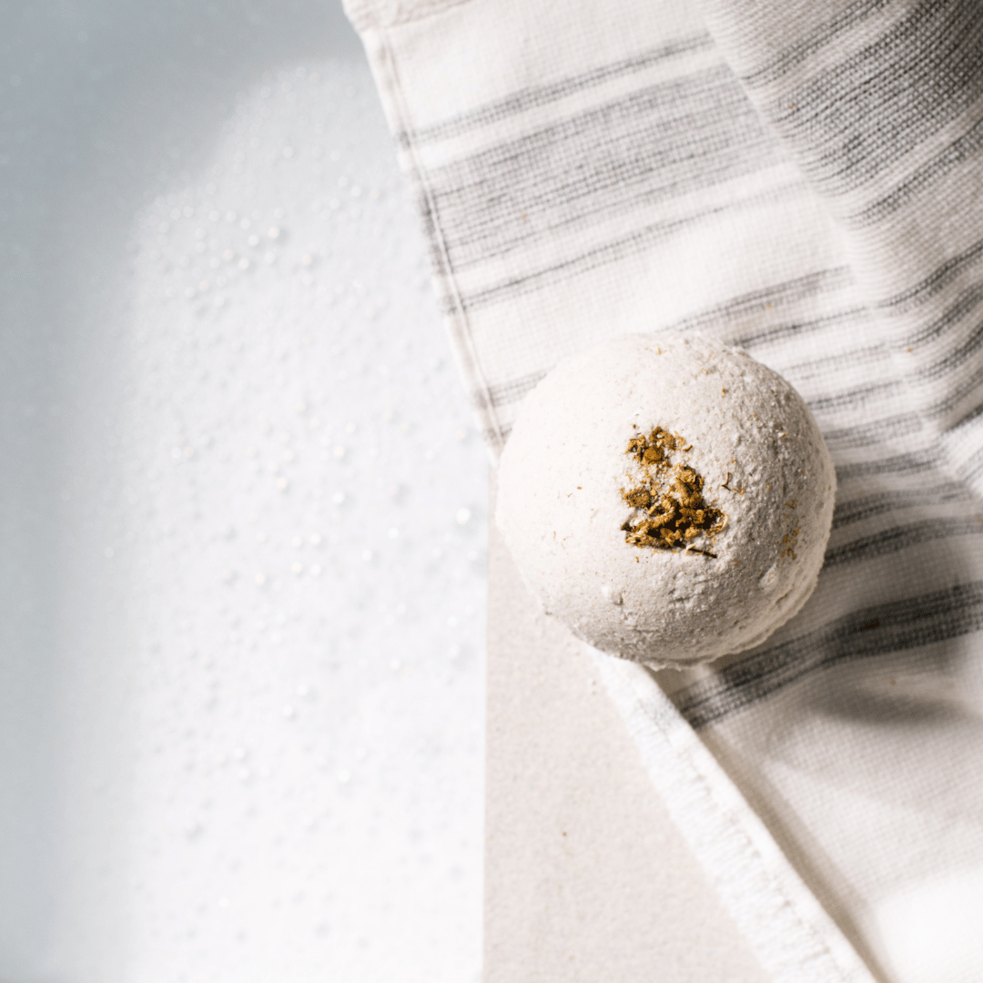 A white bath bomb with dried chamomile on top, sitting on a towel on the side of a bath tub full of water, made by Ebony & Ivory Candle Co in Lethbridge, Alberta, Canada