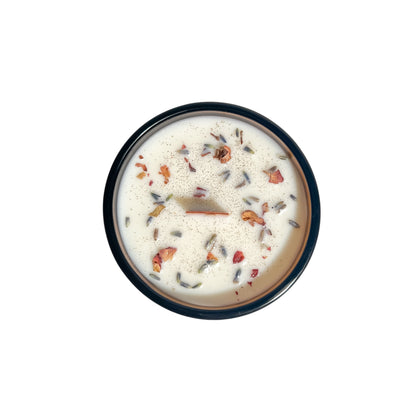 An amber jar 16oz soy wax candle with a wood wick, a gold lid, and an indigo label named "practical magic" on a white background topped with lavender buds and rose petals