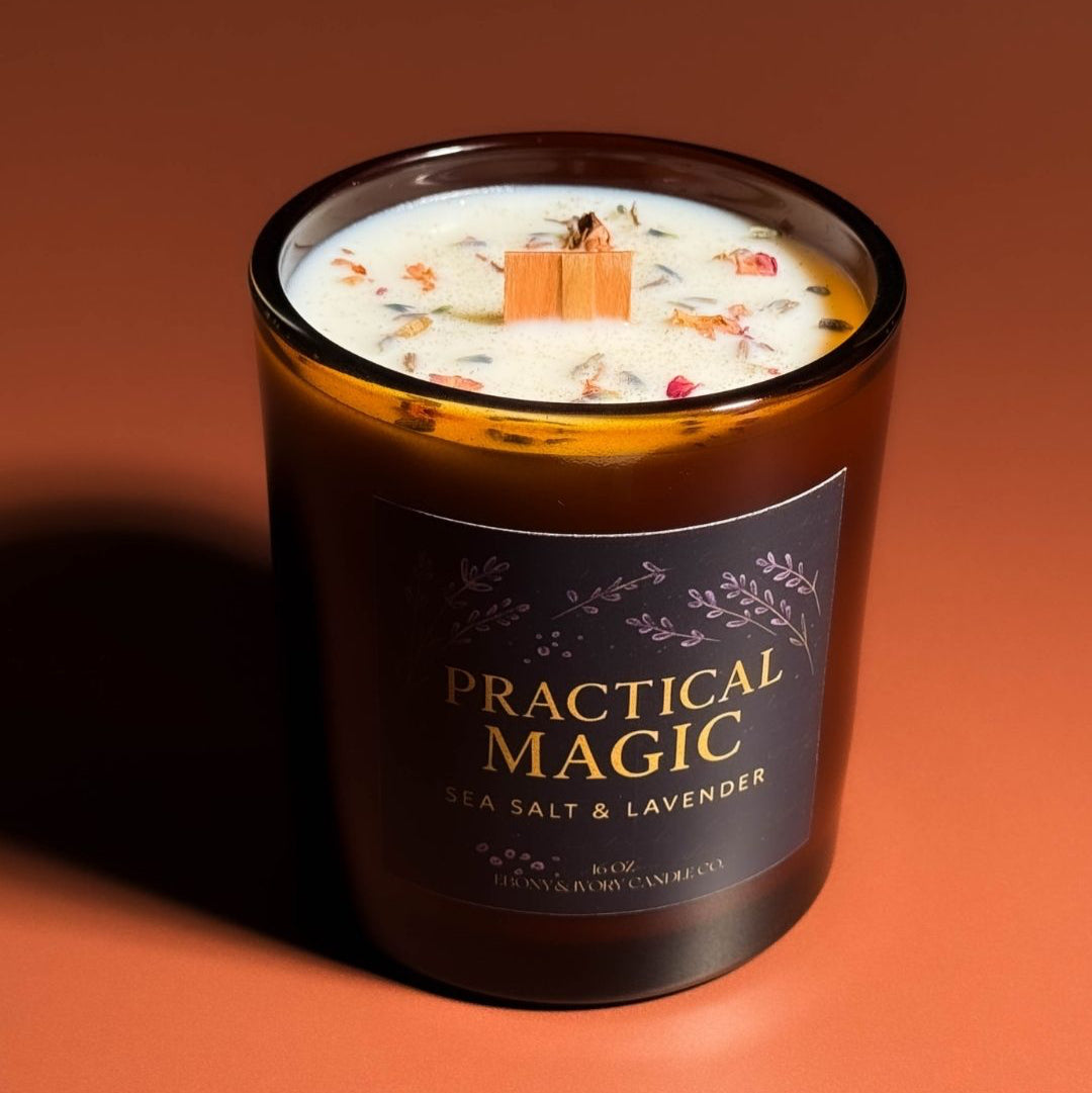 An amber jar 16oz soy wax candle with a wood wick, a gold lid, and an indigo label named "practical magic" on an amber background topped with lavender buds and rose petals