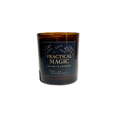 An amber jar 16oz soy wax candle with a wood wick, a gold lid, and an indigo label named "practical magic"