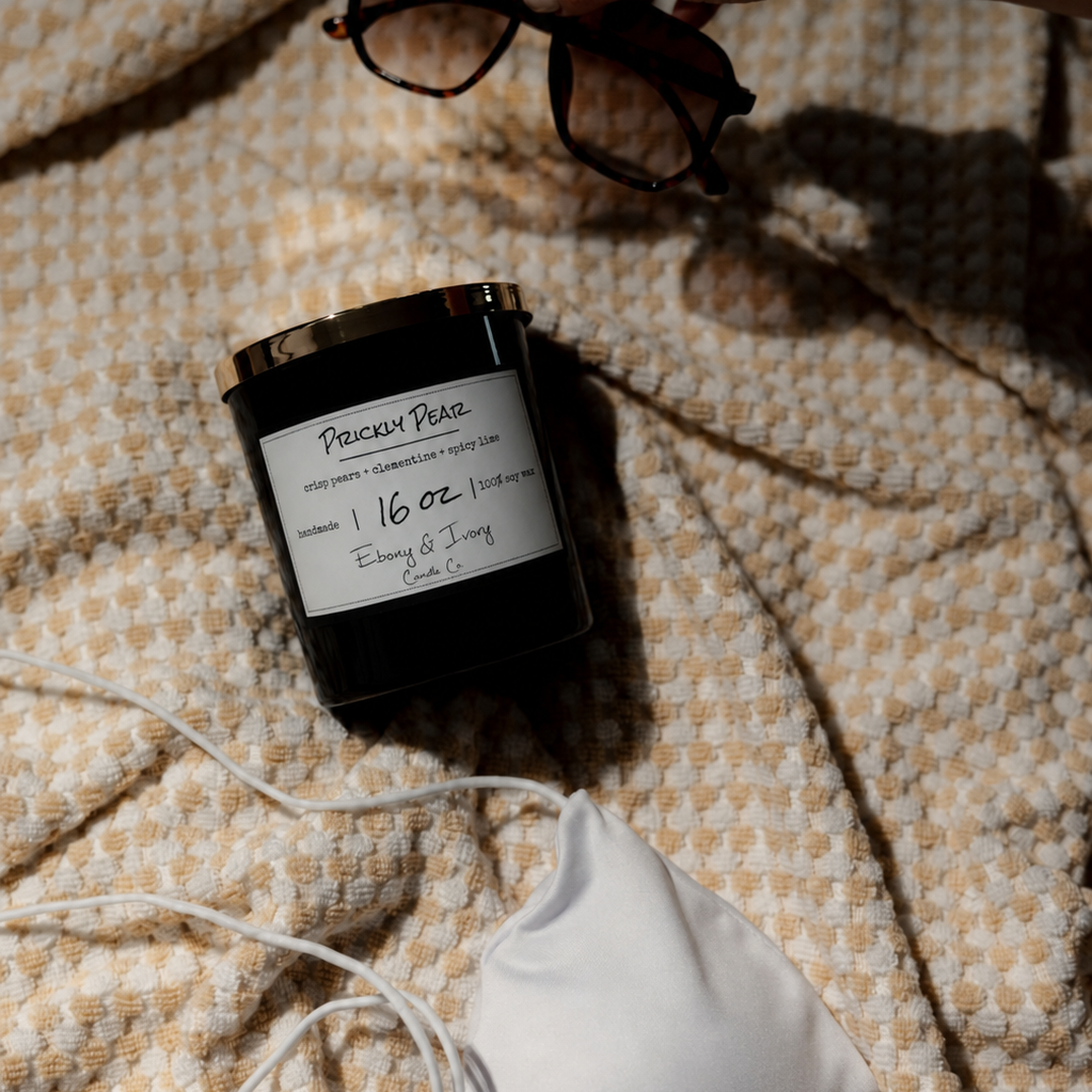 A black 16 oz soy candle with a gold lid and a white label, named Prickly Pear, laying on a beach towel with a white bikini top and sunglasses.