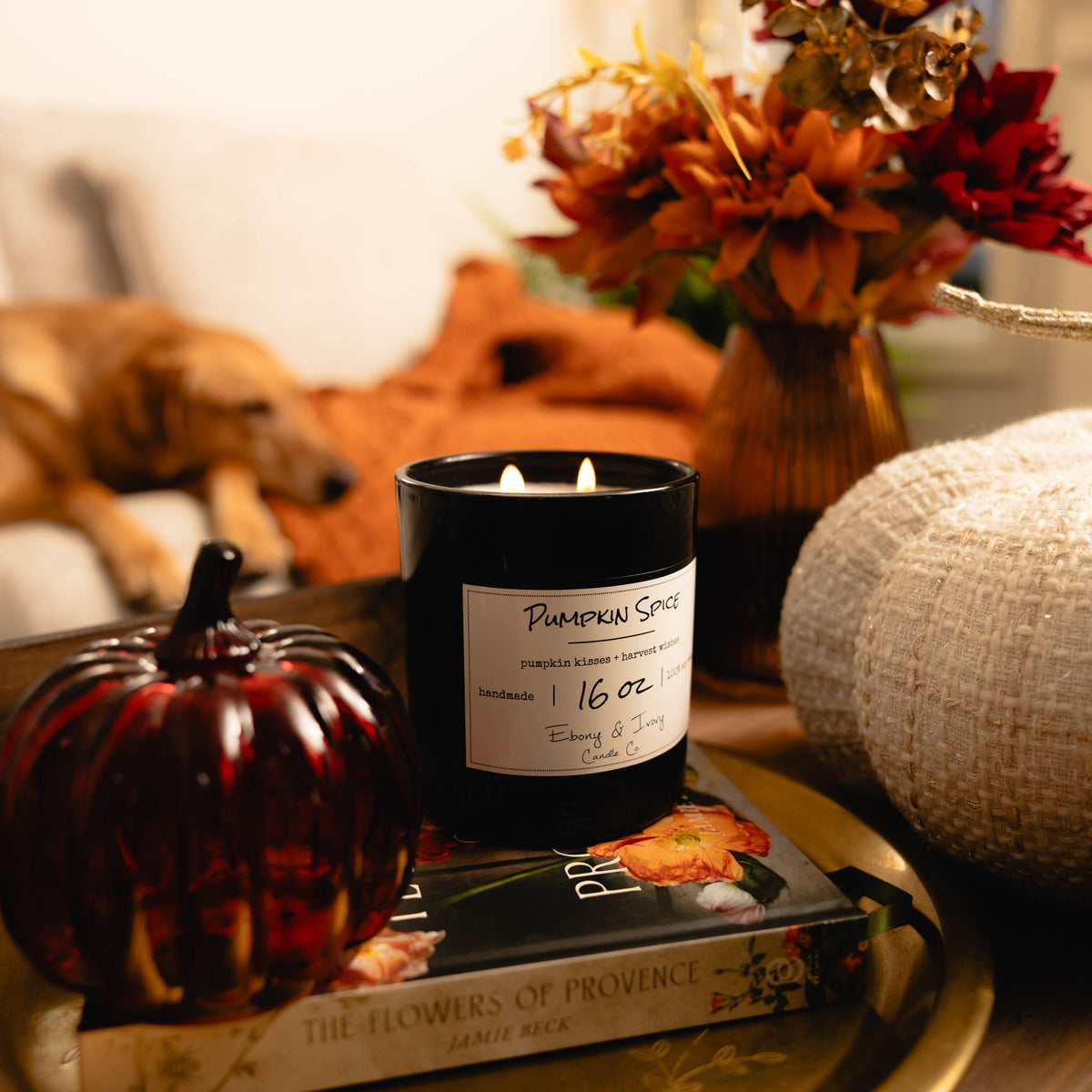 A black 16oz soy candle with a white label named "pumpkin Spice" sitting on a tabled with a book and fall decor around it.