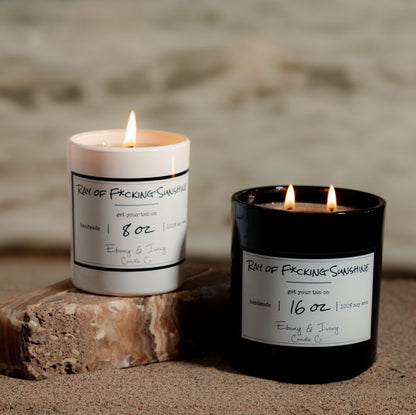 A white 8oz and a black 16oz soy candles labelled Ray of F*cking Sunshine sitting on a beach. Made in lethbridge alberta canada