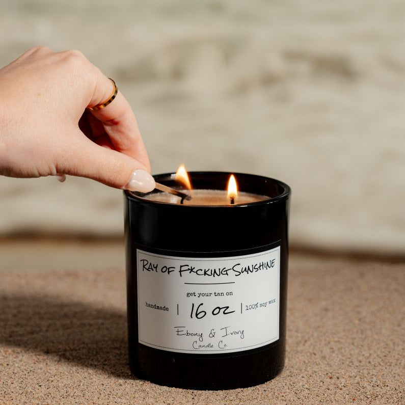 A black 16oz soy candle labelled Ray of F*cking Sunshine. Sitting on a beach, being lit by a woman. Made in lethbridge, alberta, canada