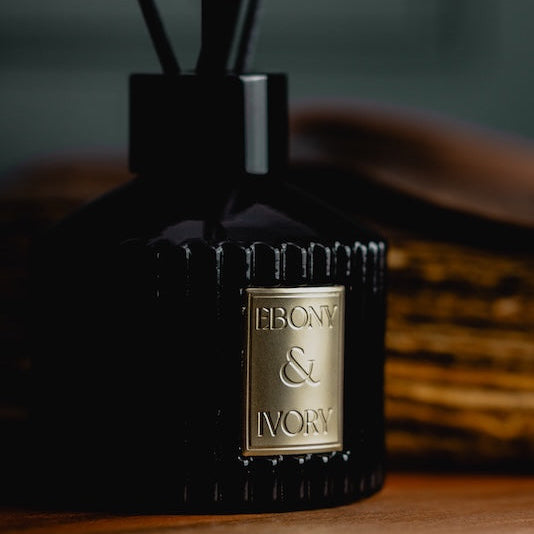 A black 100ml ribbed luxx reed diffuser with black diffuser reeds and a gold Ebony & Ivory sticker, sitting on a wooden table with a leather journal behind it and a dark green wall, made by Ebony & Ivory Candle Co in Lethbridge, Alberta, Canada