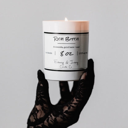 A white jar 8oz soy candle with a white label named Rich Bitch, on a white background, being held by a hand with a luxurious lacey black glove, made by Ebony & Ivory Candle Co in Lethbridge, Alberta, Canada