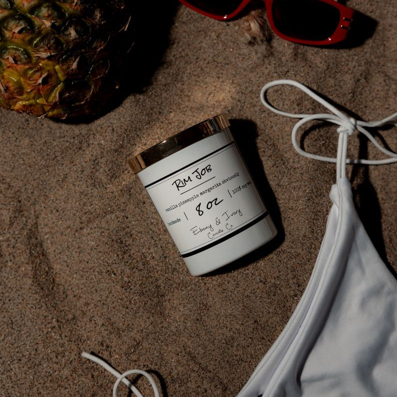 A white 8oz soy candle with a gold lid, labelled Rim Job, laying on the beach. Made in Lethbridge, alberta, canada