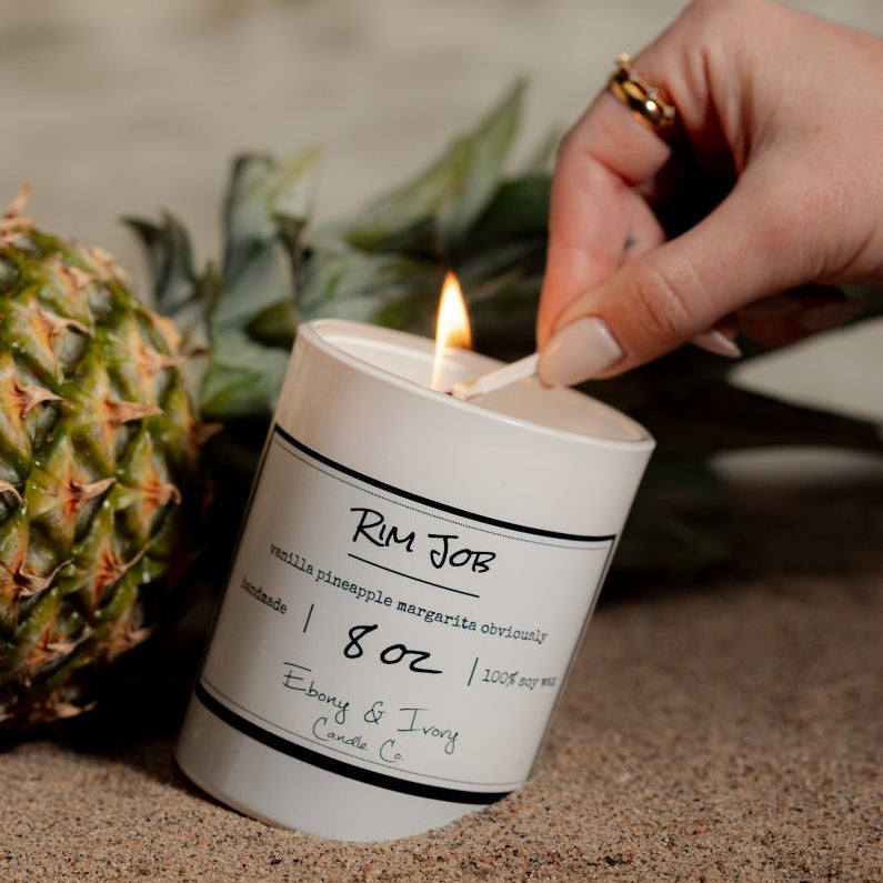 A white 8oz soy candle buried in the sand on the beach with a hand lighting it. The candle is labelled Rim Job. Made in Lethbridge, Alberta, Canada