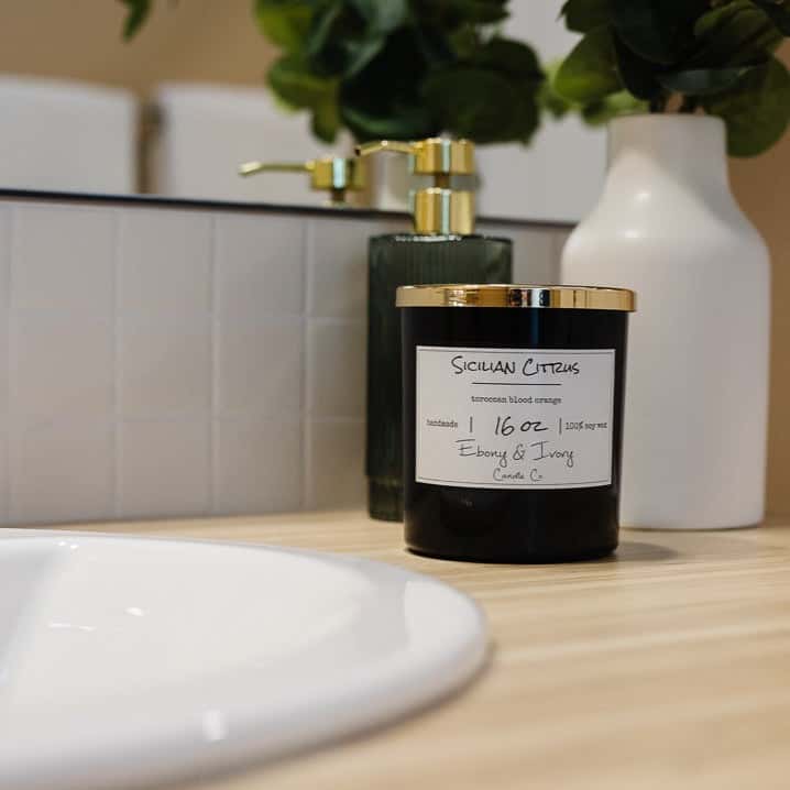 A black jar 16oz soy candle with a gold lid and a white label named Sicilian Citrus, on a light wood bathroom counter, with an elegant green soap pump and a vase of flowers behind it, made by Ebony & Ivory Candle Co in Lethbridge, Alberta, Canada