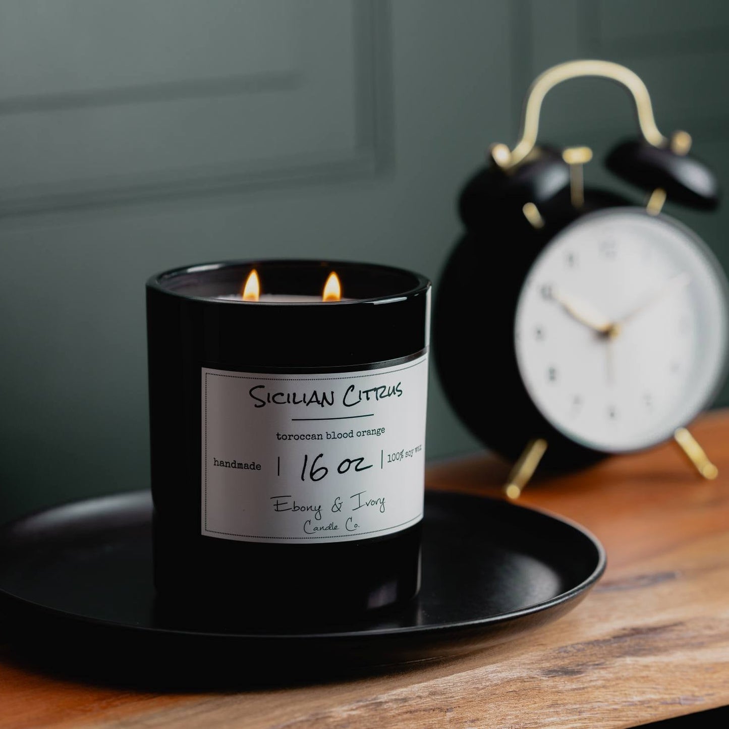 A black jar 16oz soy candle with a gold lid and a white label named Sicilian Citrus, on a black tray on top of a wooden table, with a black alarm clock behind it, and a moody dark green wall. made by Ebony & Ivory Candle Co in Lethbridge, Alberta, Canada.
