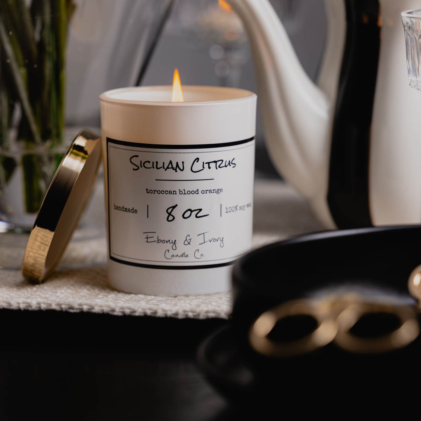 A white jar 8oz soy candle with a gold lid and a white label named Sicilian Citrus, on a black dining room table, with a tea pot and wine glasses behind it, made by Ebony & Ivory Candle Co in Lethbridge, Alberta, Canada
