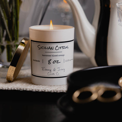 A white jar 8oz soy candle with a gold lid and a white label named Sicilian Citrus, on a black dining room table, with a tea pot and wine glasses behind it, made by Ebony & Ivory Candle Co in Lethbridge, Alberta, Canada