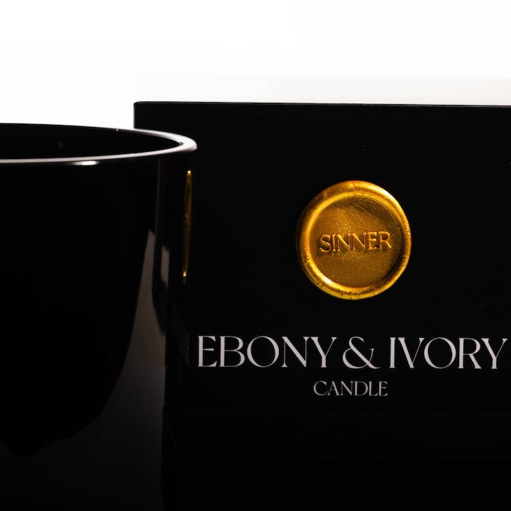 A close up of a black 18oz jar coconut soy candle with a white Ebony & Ivory on the front next to a black candle box with a gold label named Sinner, on a white background, made by Ebony & Ivory Candle Co in Lethbridge, Alberta, Canada