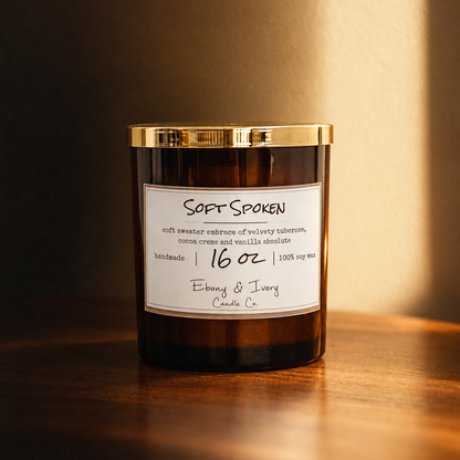 An amber 16oz glass jar soy candle with a gold lid and a white label named "soft spoken" on a wooden table and soft autumn lighting