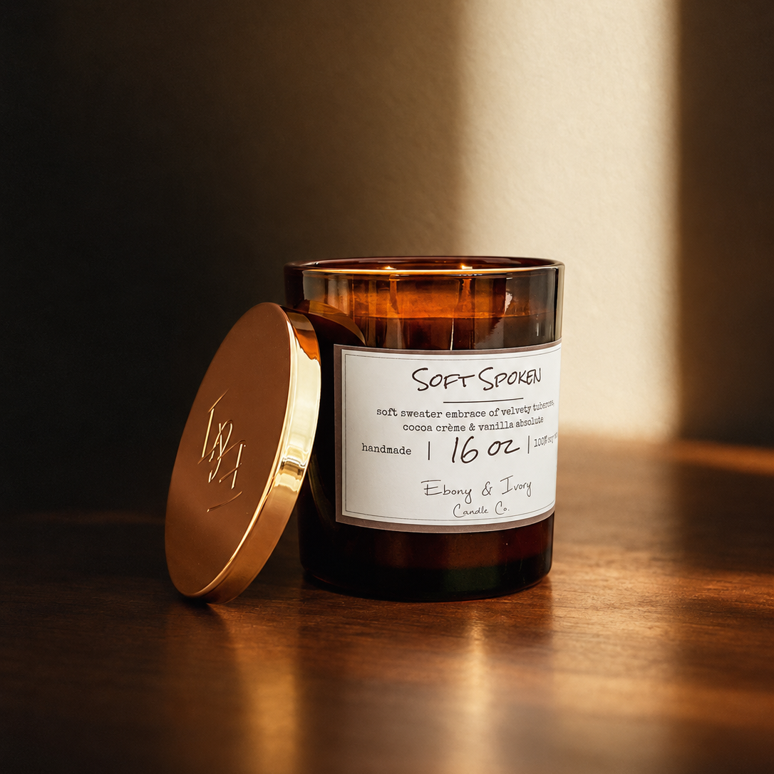 A 16oz amber glass jar soy candle with a gold lid and two cotton wicks and a white label named "Soft Spoken" sitting on a wooden table with soft autumn lighting.