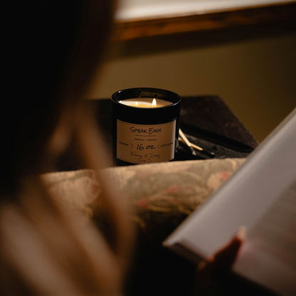 A black 16oz soy candle with two lit cotton wicks on a wooden side table next to someone reading. The candle has a white label named "speak easy"