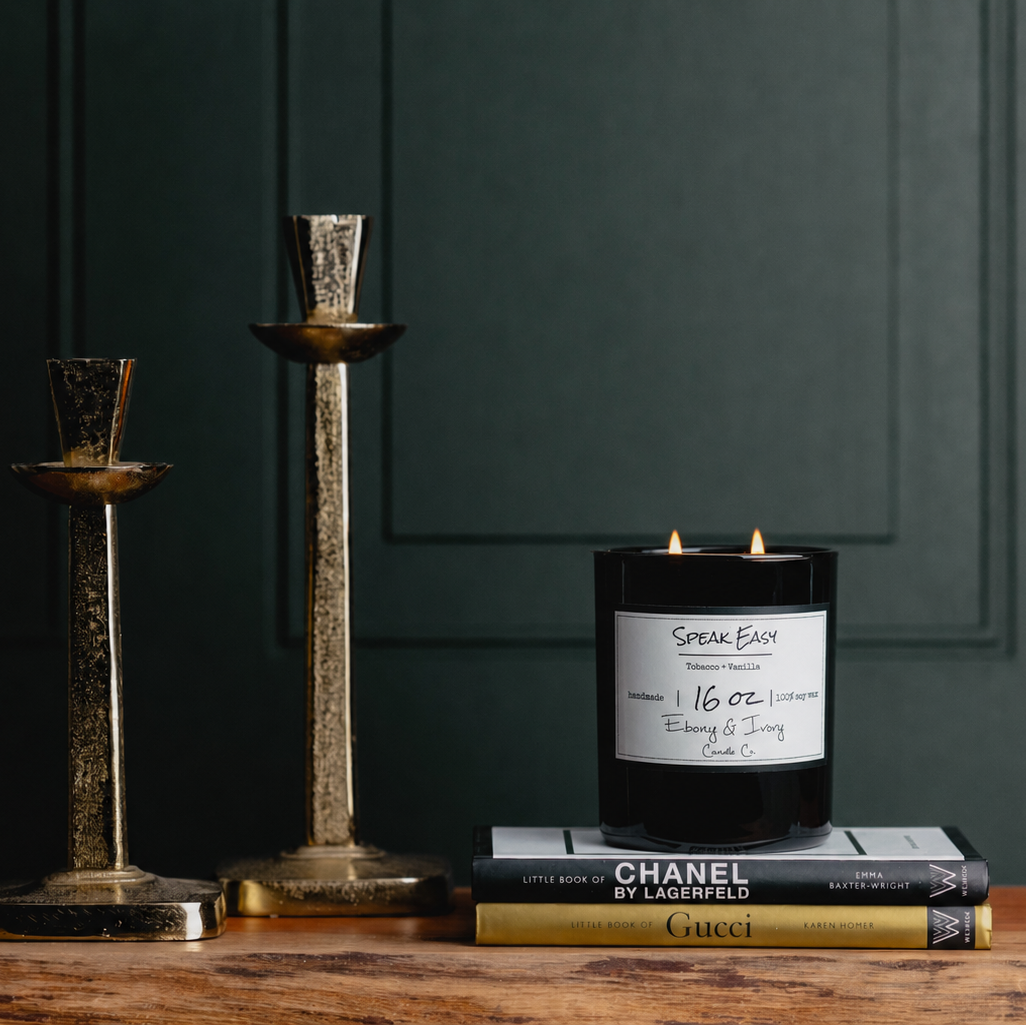 A black 16oz soy candle with two lit cotton wicks sitting on a stack of designer books, next to two gold candle stick holders. The candle has a white label named "Speak Easy"