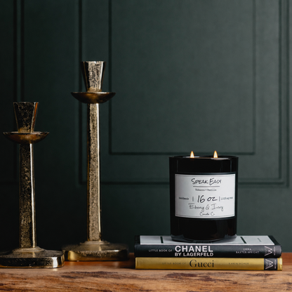 A black 16oz soy candle with two lit cotton wicks sitting on a stack of designer books, next to two gold candle stick holders. The candle has a white label named "Speak Easy"