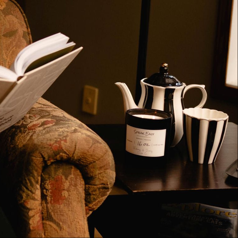 A black 16oz soy candle with 2 lit cotton wicks and a white label named "Speak Easy" with a black and white striped tea pot and mug, on a wooden side table next to a cozy chair and someone reading.