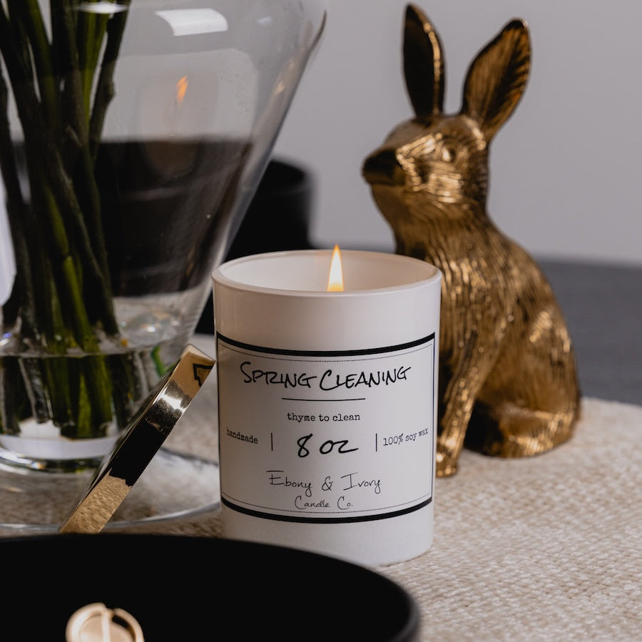 Candle with a label on a surface next to a gold rabbit figurine and a vase with greenery.