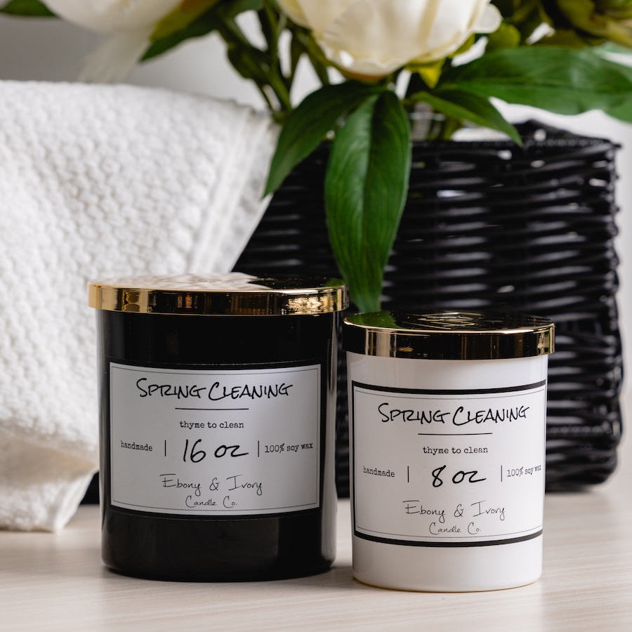 Two candles labeled 'Spring Cleaning
' with a vase of white flowers in the background.