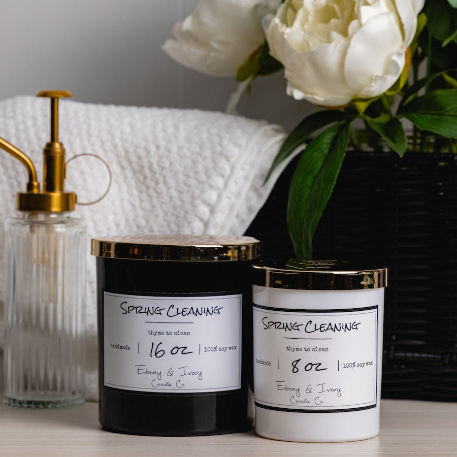 Two candles labeled 'Spring Cleaning' on a surface with flowers and a towel in the background.