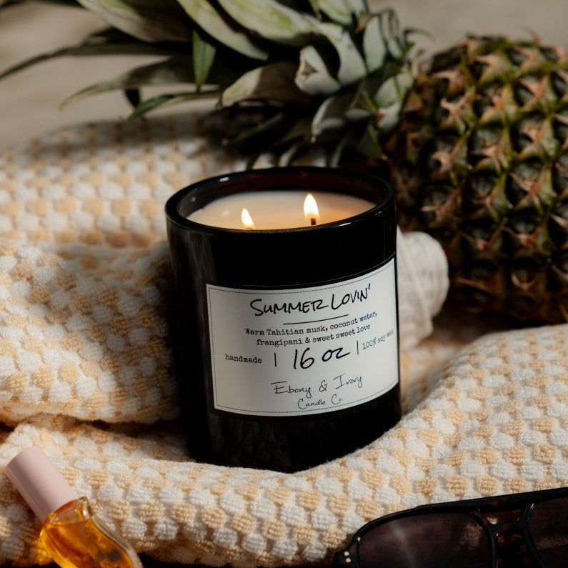 A black 16oz soy candle labelled Summer Lovin', sitting on a beach towel with sunglasses and a pineapple. Made in Lethbridge Alberta Canada