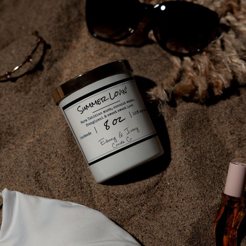 A white 8oz soy candle with a gold lid, labelled Summer Lovin, laying on a back with a white bikini and sunglasses. Made in Lethbridge, Alberta, Canada