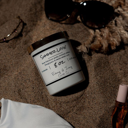 A white 8oz soy candle with a gold lid, labelled Summer Lovin, laying on a back with a white bikini and sunglasses. Made in Lethbridge, Alberta, Canada