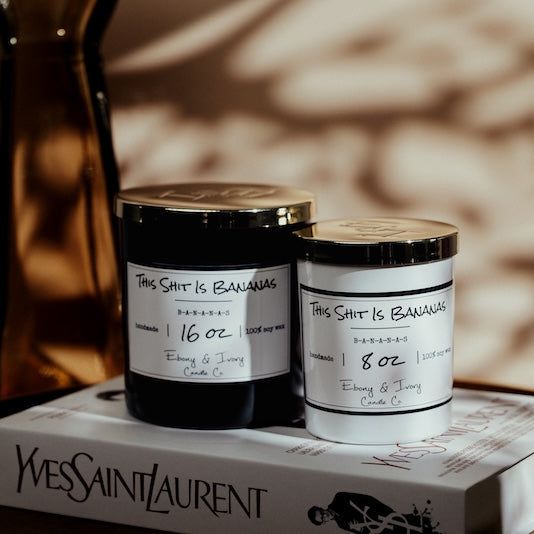 Two candles with labels on a Yves Saint Laurent box against a blurred background. Yves saint Laurent book decor with Canadas soy candle. Banana scented soy candle Canada.