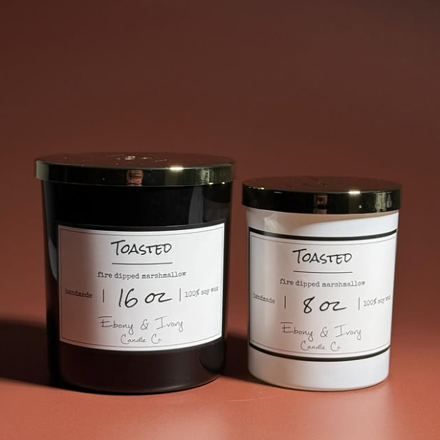 A black jar 16oz soy candle with a gold lid and a white label named Toasted, next to a white 8oz jar soy candle with a gold lid and a white label named Toasted. on a cozy rusty red background, made by Ebony & Ivory Candle Co in Lethbridge, Alberta, Canada