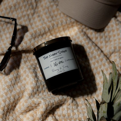 A black 16 ounce soy candle with a gold lid and a white label named "Two Night Stand" Laying on a white and orange beach towel with sunglasses, a baseball cap, and a pineapple. 