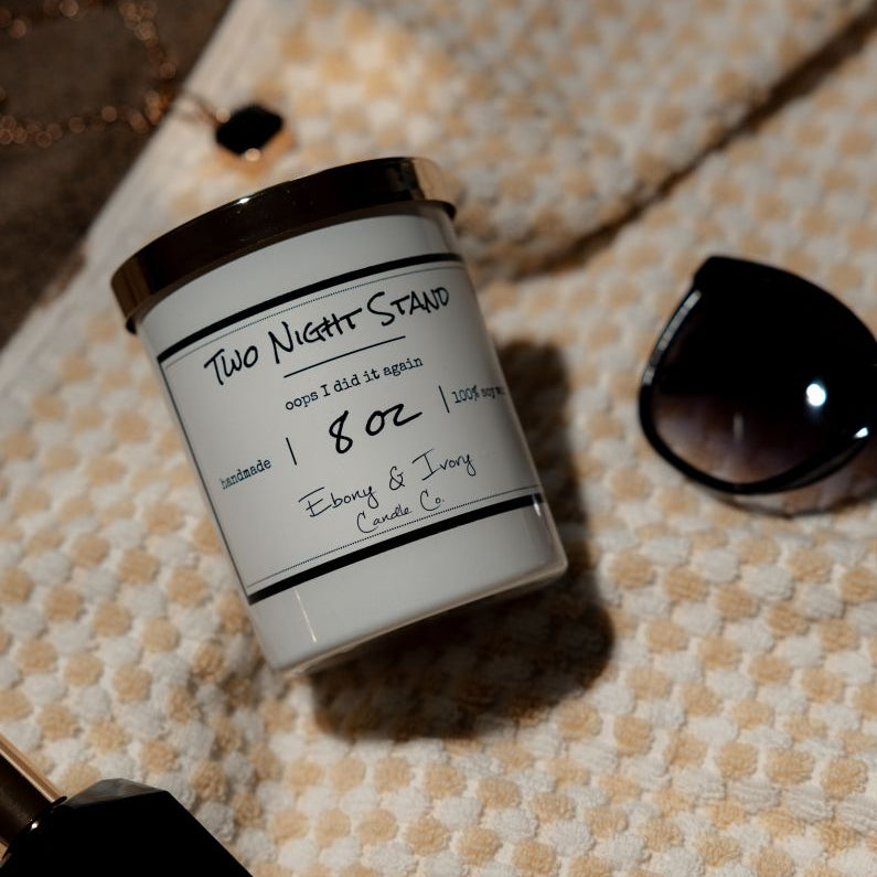 A white 8 ounce soy candle with a gold lid and a white label named "Two Night Stand" laying on a white and orange beach towel with sunglasses, a gold necklace, and a perfume made by Ebony & Ivory Candle Co.