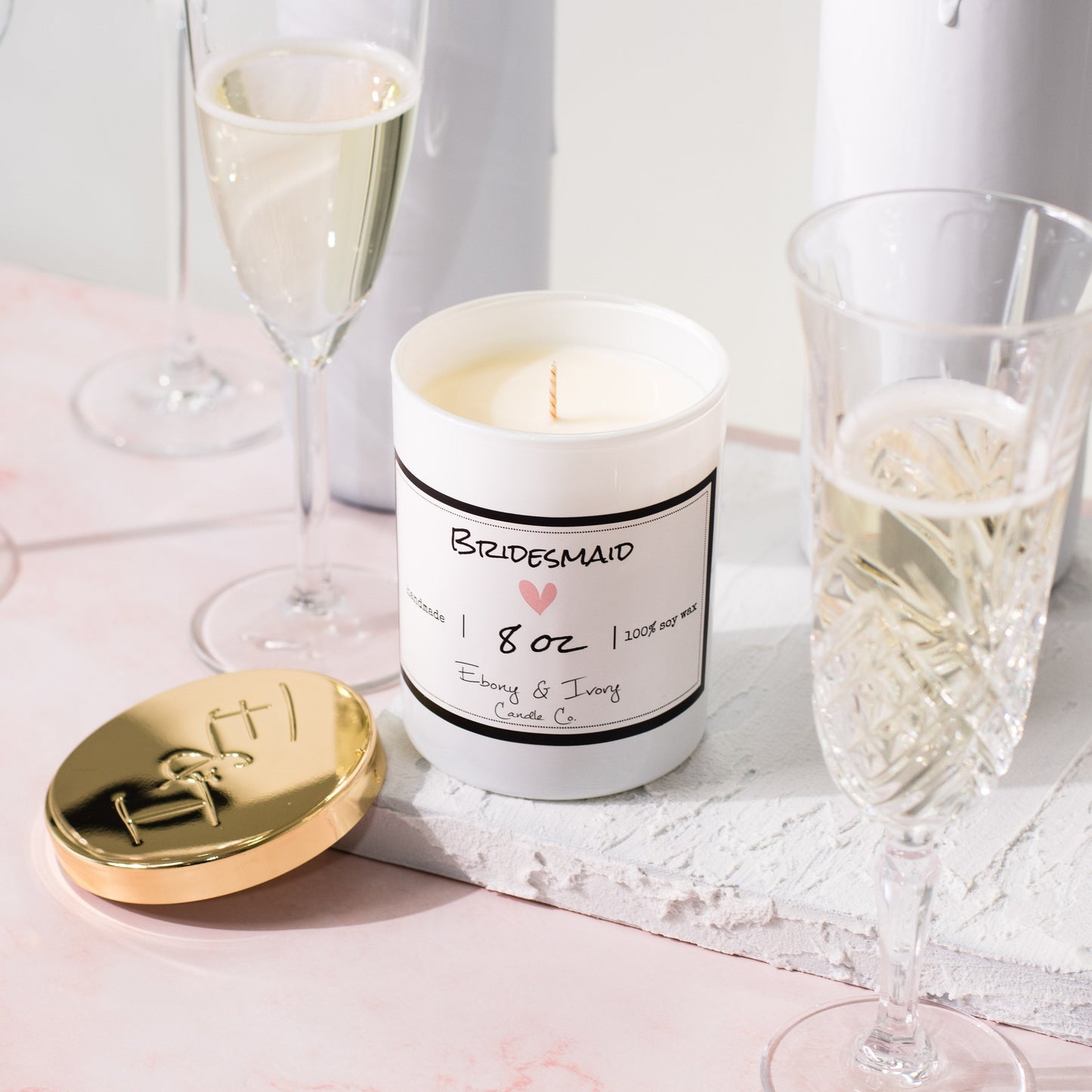 A white jar 8oz soy candle with a gold lid and a white label named Bridesmaid, on a white tray on a wedding table, with champagne glasses, made by Ebony & Ivory Candle Co in Lethbridge, Alberta, Canada