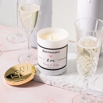 A white jar 8oz soy candle with a gold lid and a white label named Bridesmaid, on a white tray on a wedding table, with champagne glasses, made by Ebony & Ivory Candle Co in Lethbridge, Alberta, Canada