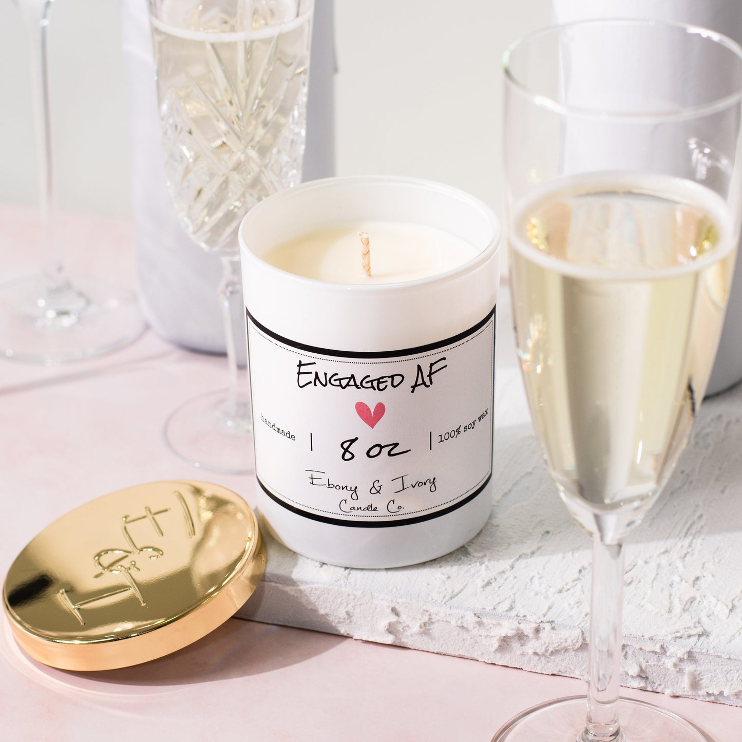 A white jar 8oz soy candle with a gold lid and a white label named Engaged AF, on a white tray on a wedding table, with champagne glasses, made by Ebony & Ivory Candle Co in Lethbridge, Alberta, Canada