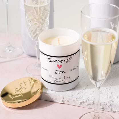 A white jar 8oz soy candle with a gold lid and a white label named Engaged AF, on a white tray on a wedding table, with champagne glasses, made by Ebony & Ivory Candle Co in Lethbridge, Alberta, Canada