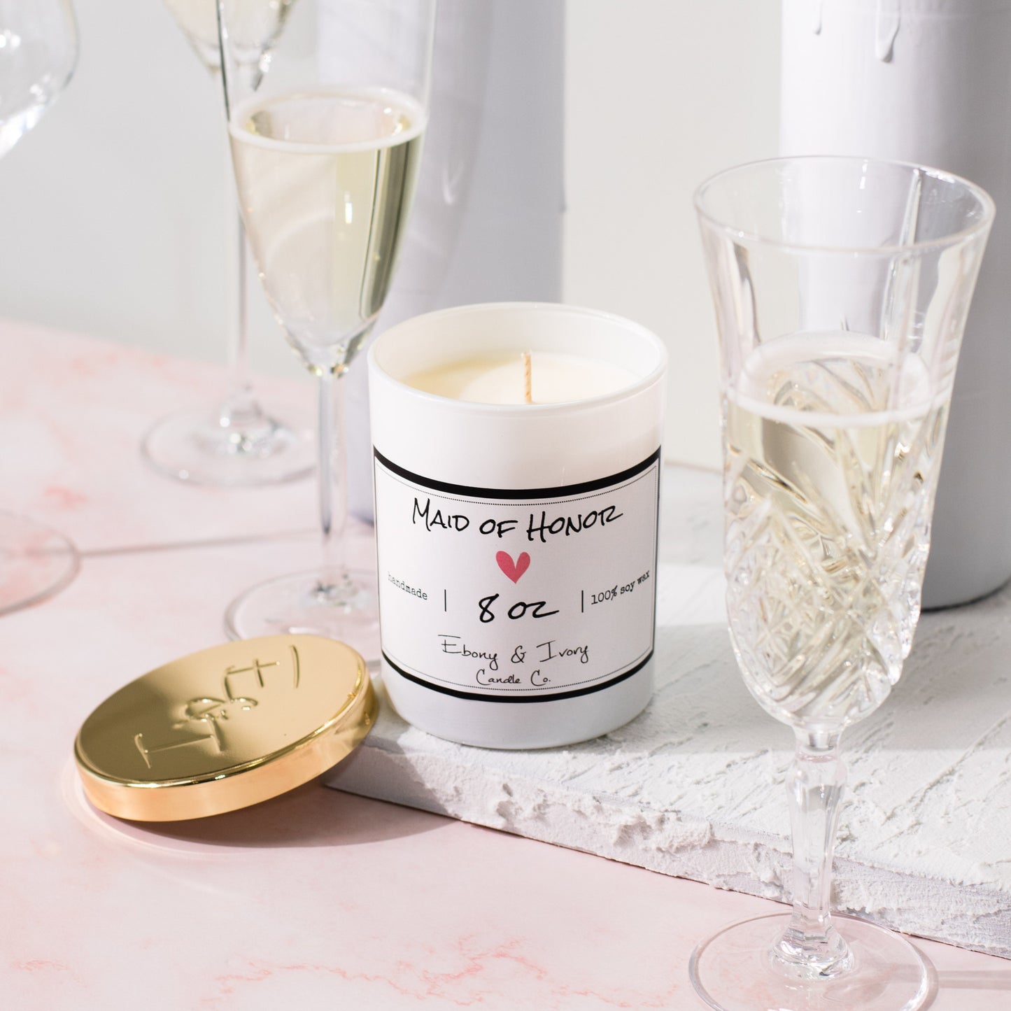 A white jar 8oz soy candle with a gold lid and a white label named Maid of Honor, on a white tray on a wedding table, with champagne glasses, made by Ebony & Ivory Candle Co in Lethbridge, Alberta, Canada