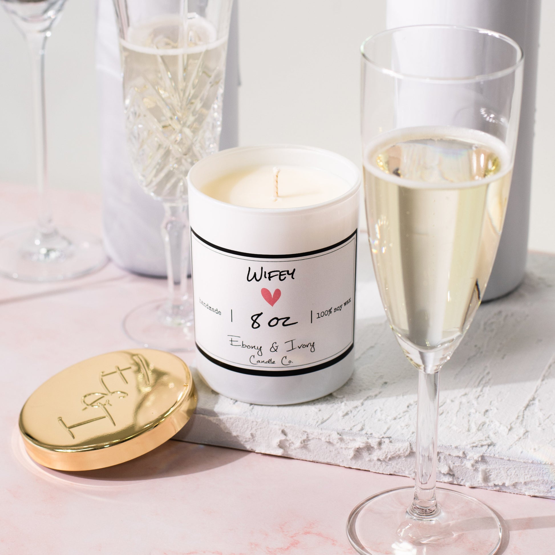 A white jar 8oz soy candle with a gold lid and a white label named Wifey, on a white tray on a wedding table, with champagne glasses, made by Ebony & Ivory Candle Co in Lethbridge, Alberta, Canada
